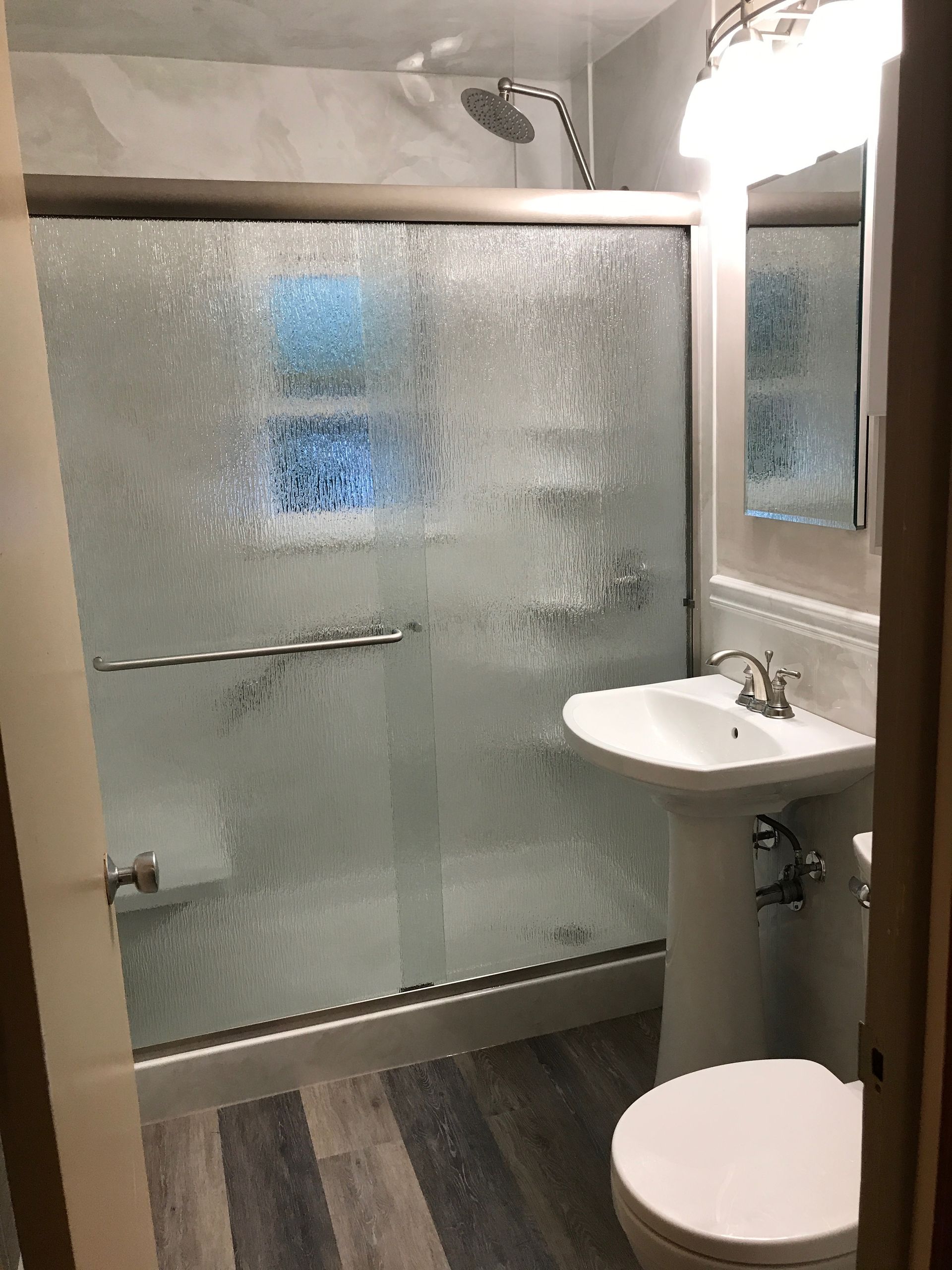 Bathroom with shower, pedestal sink, and toilet; gray wood-look floor, frosted glass shower door, and light gray walls.
