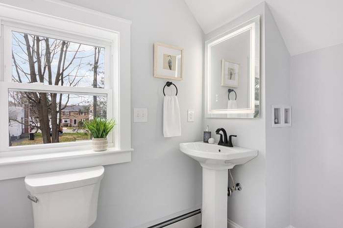 Bathroom with white toilet, pedestal sink, window, mirror, and light blue walls.