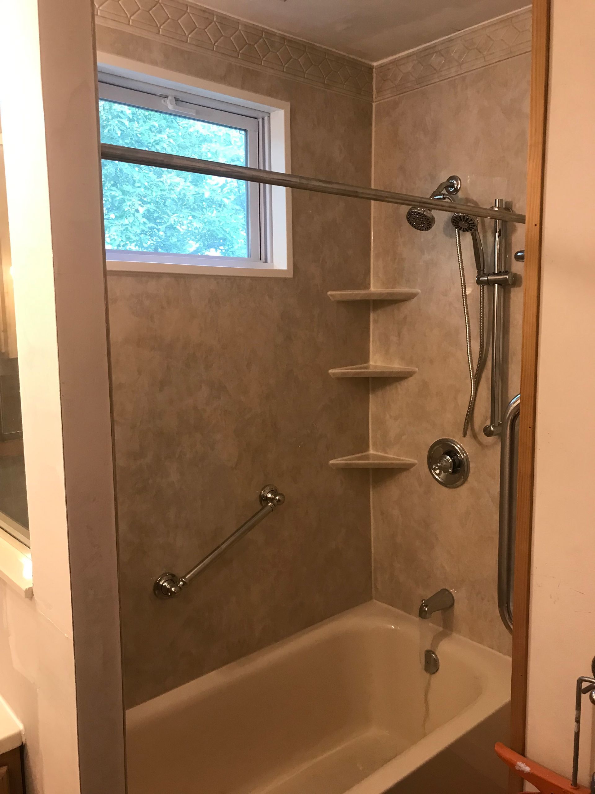 Bathtub with tan walls, shelves, grab bar, and shower head near a window.