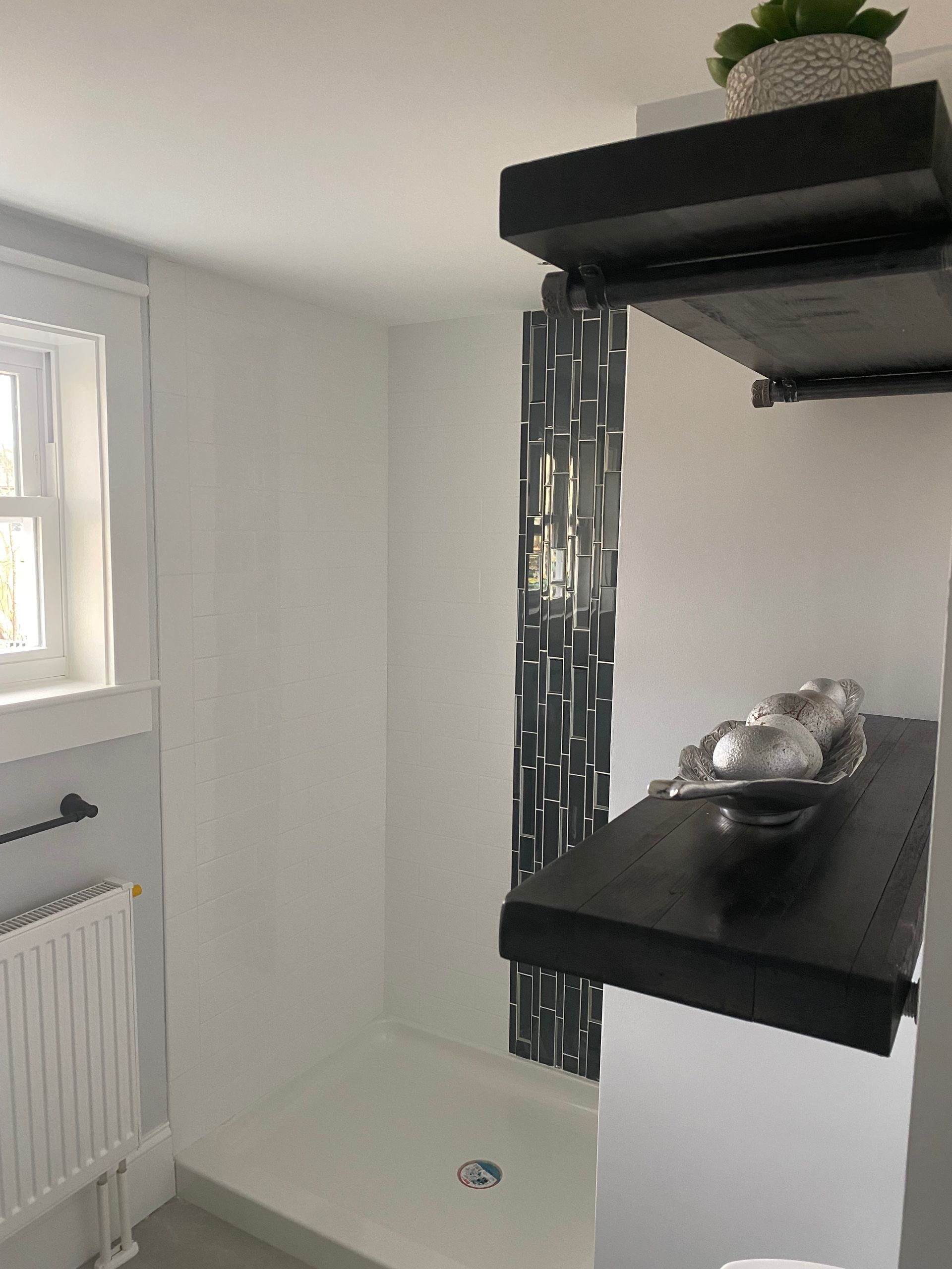 Bathroom with white walls, shower with black tile, shelves with decorations, and a window.