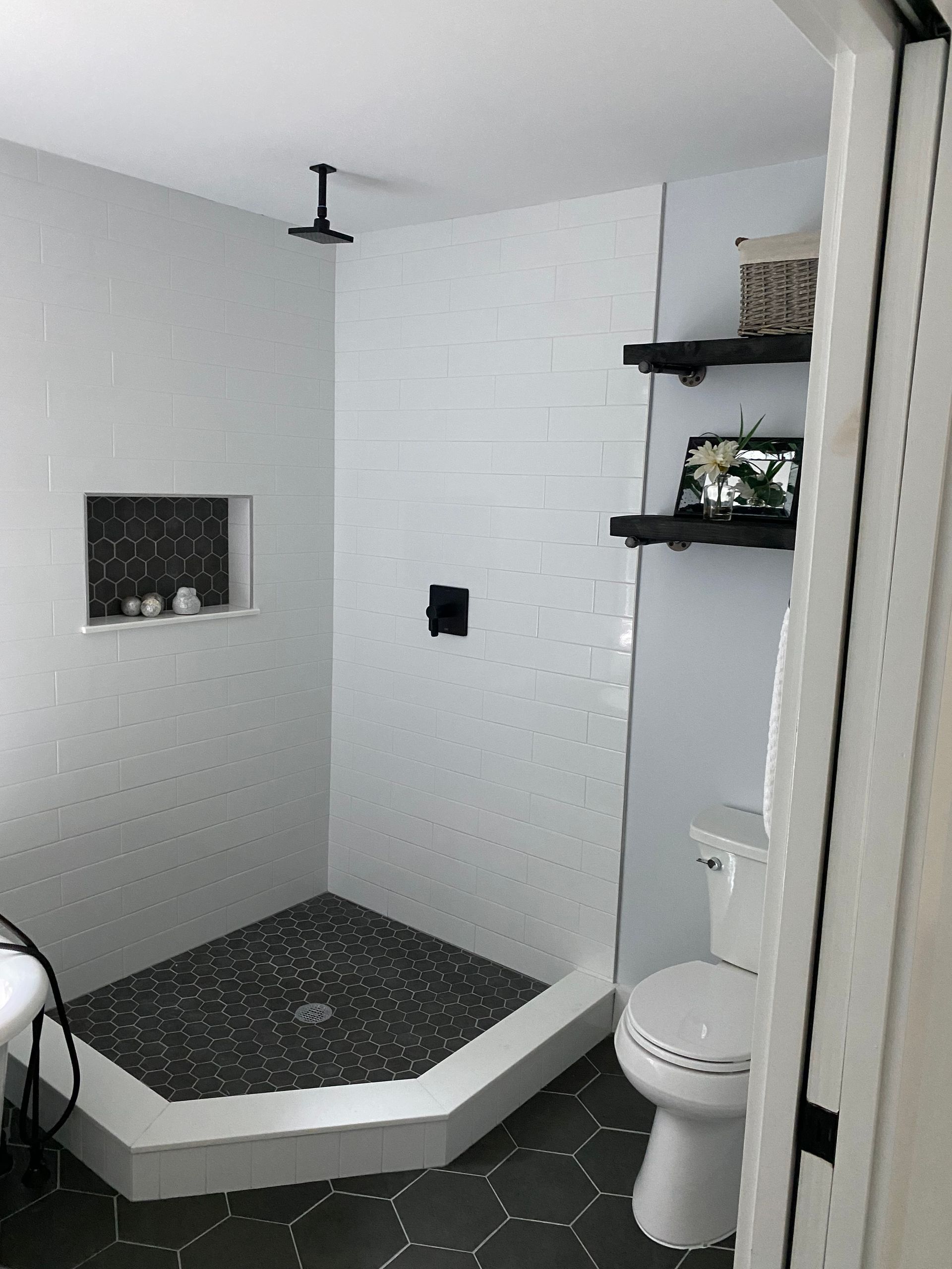 Modern bathroom with white and black tile shower, toilet, and black shelves with decor.