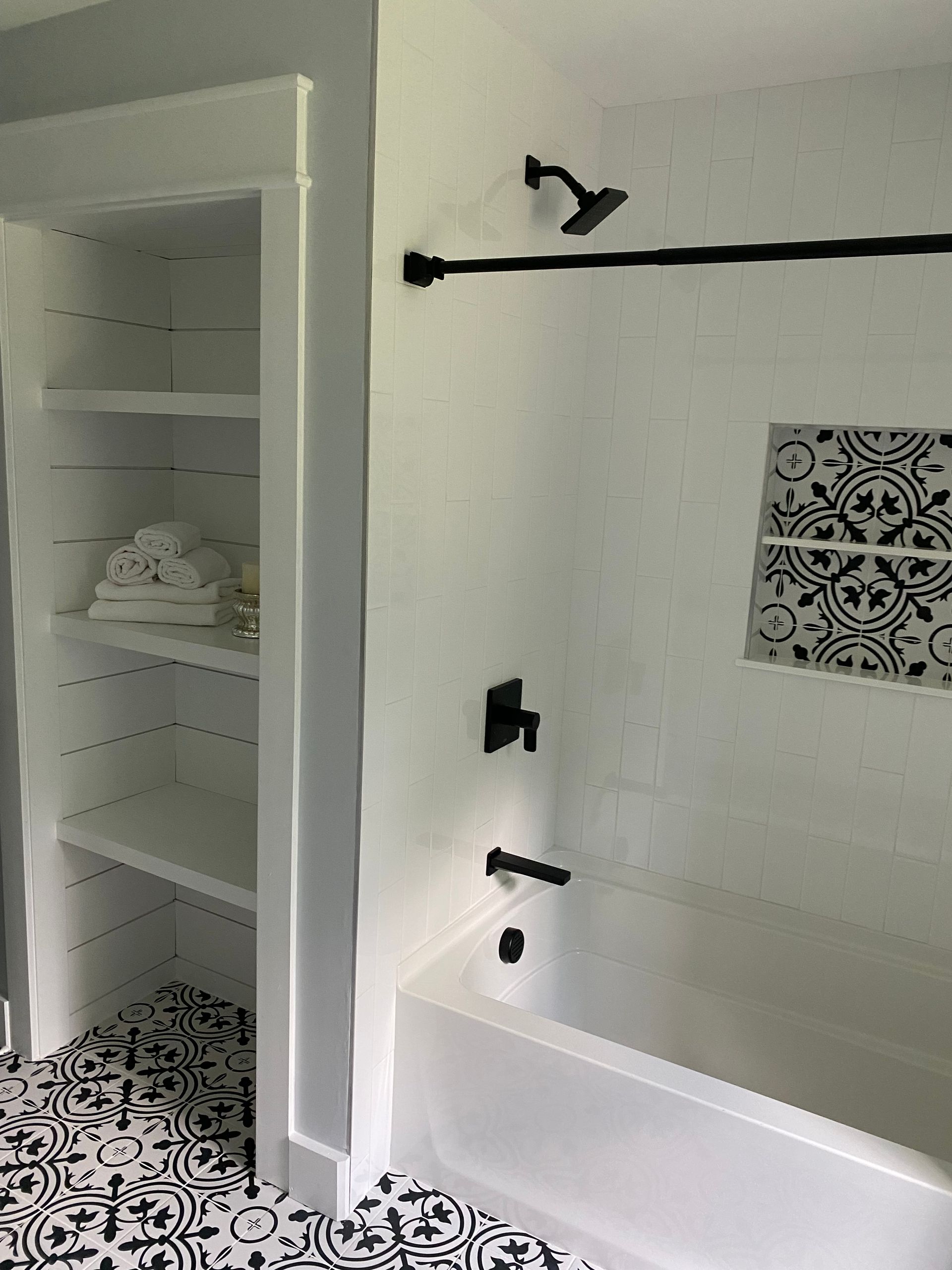 Bathroom with built-in shelves holding towels, black and white tile floor, and black fixtures over the white tub.