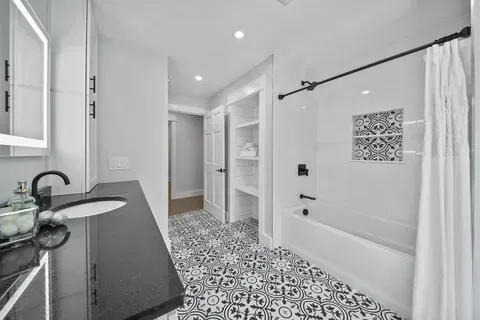 Modern white bathroom with black accents. Features vanity, tub, patterned floor, and closet.