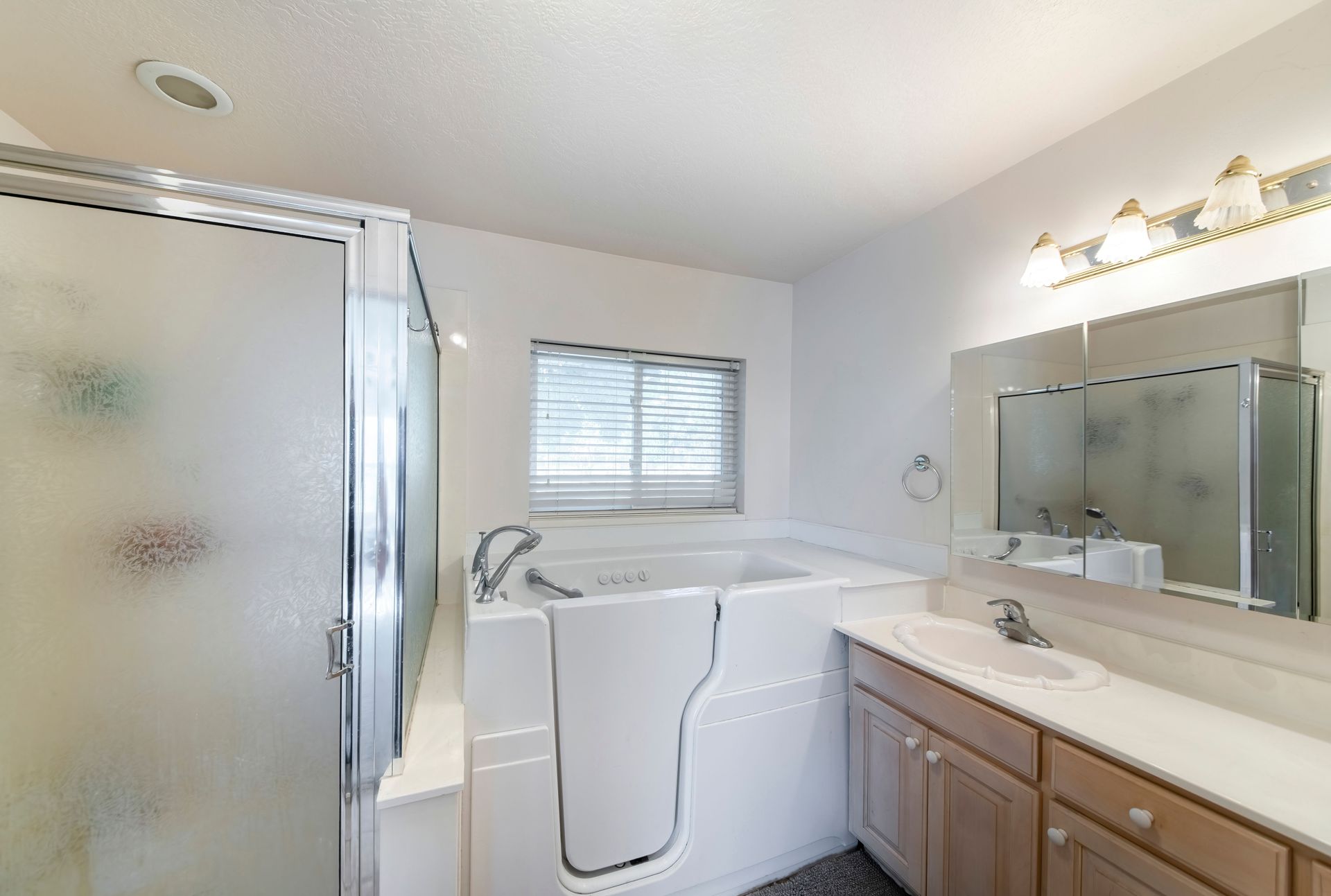 Bathroom with walk-in tub and shower. White walls, wood vanity. Window with blinds.