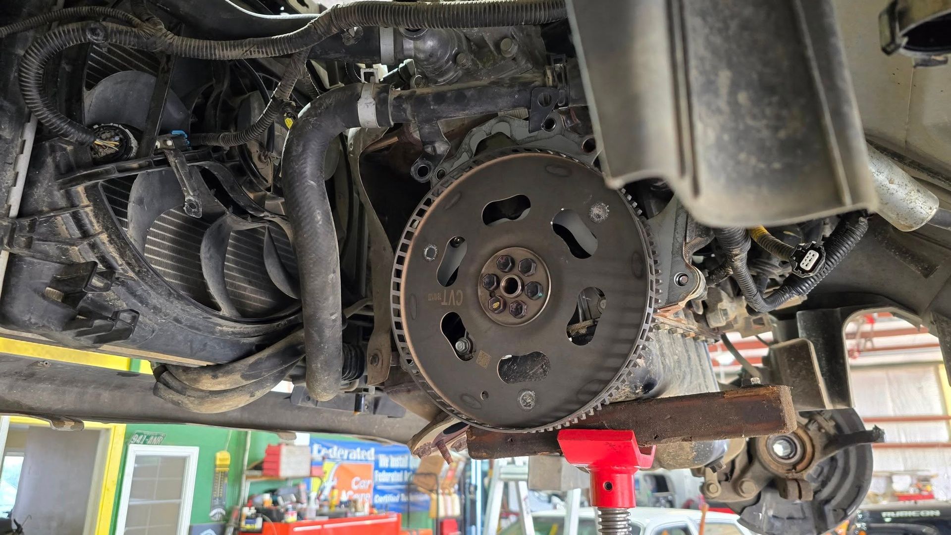 Underside view of a vehicle engine, revealing the flywheel, cooling fan, and support jack.