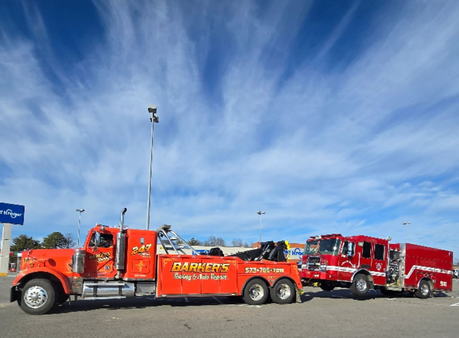 Orange tow truck towing a red fire engine in a parking lot under a blue, cloudy sky.