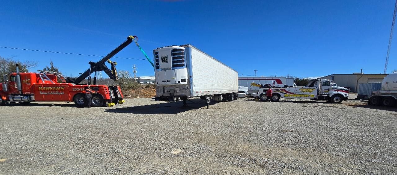 A tow truck is lifting a refrigerated trailer on a gravel lot, bright blue sky in background.