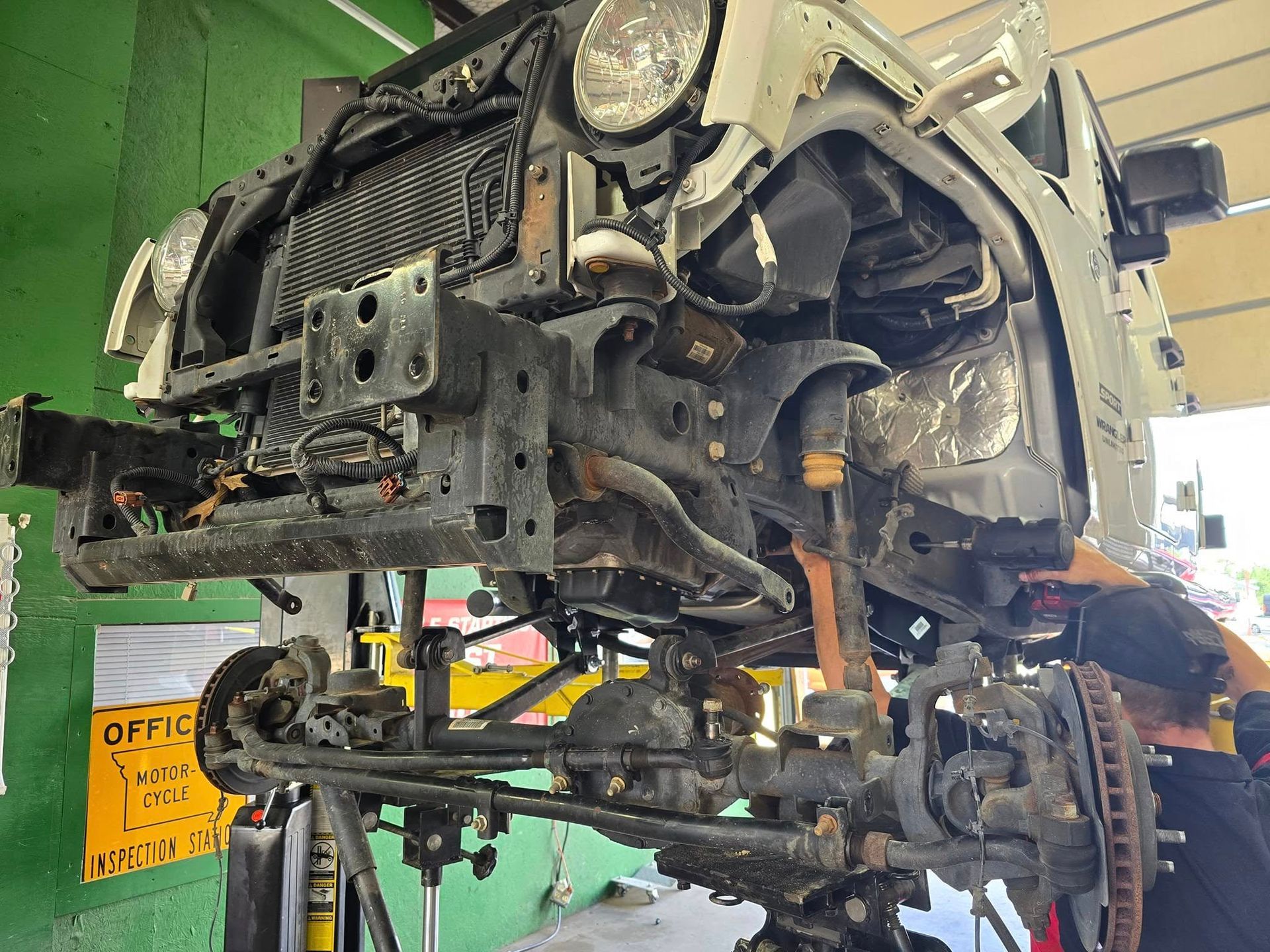 A white Jeep in a shop, front end disassembled, raised on a lift. A mechanic works on the axle.