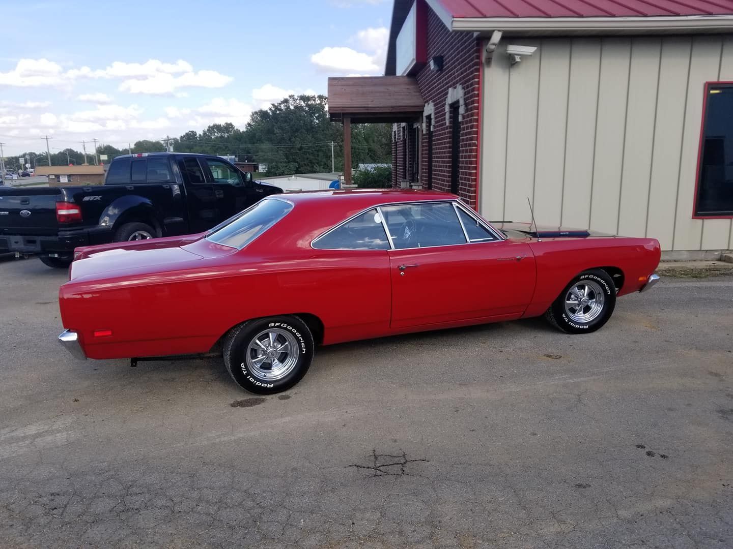 Red classic car parked outside a building; black pickup truck in the background.