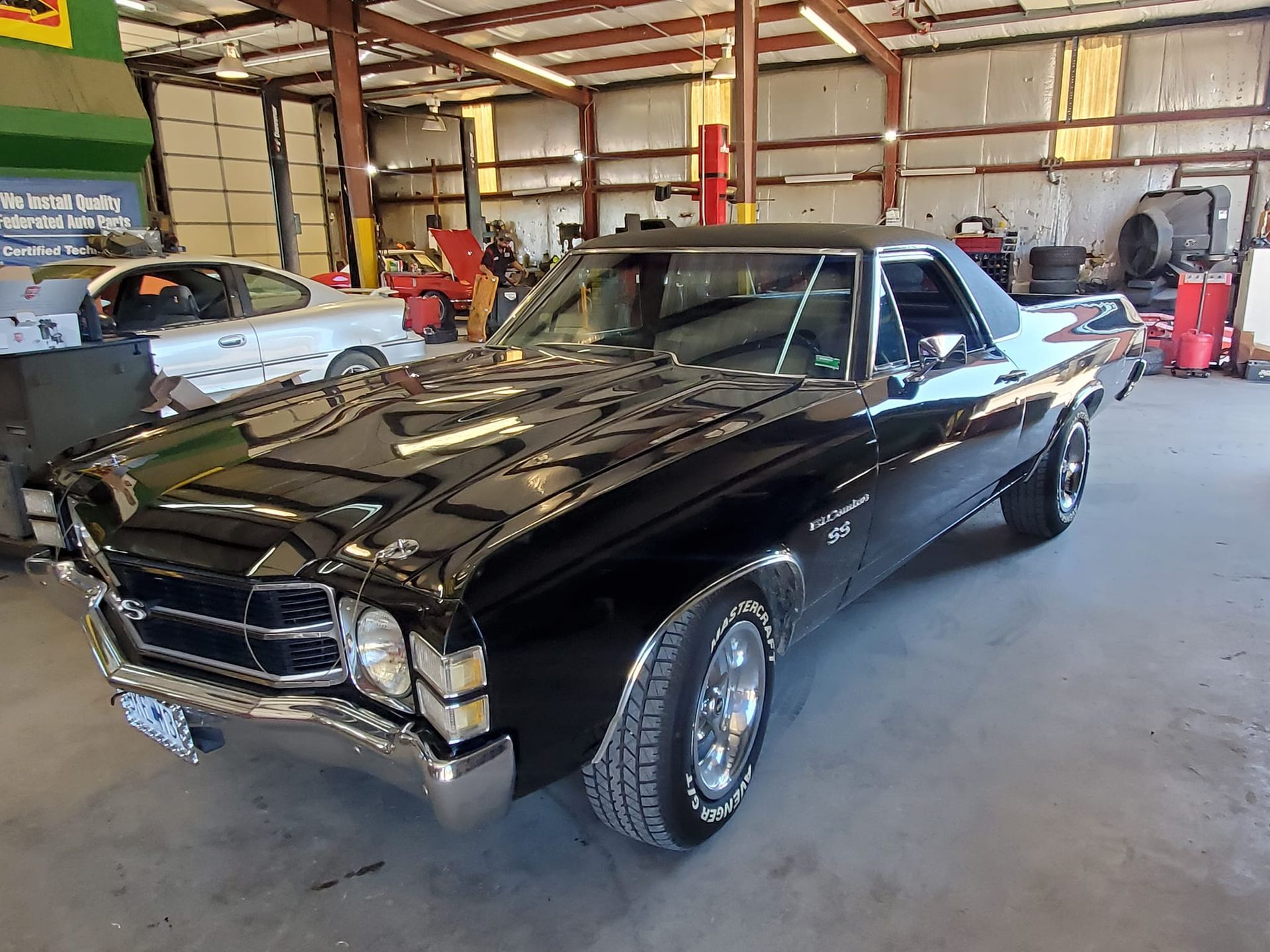 Black 1970s Chevrolet El Camino in a repair shop, chrome accents, shiny paint, with another car in the background.