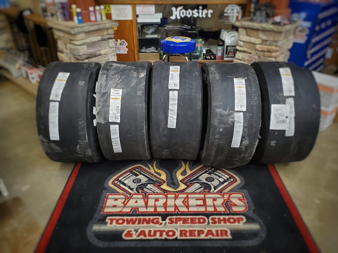 Five racing tires displayed on a Barker's Towing & Auto Repair mat.