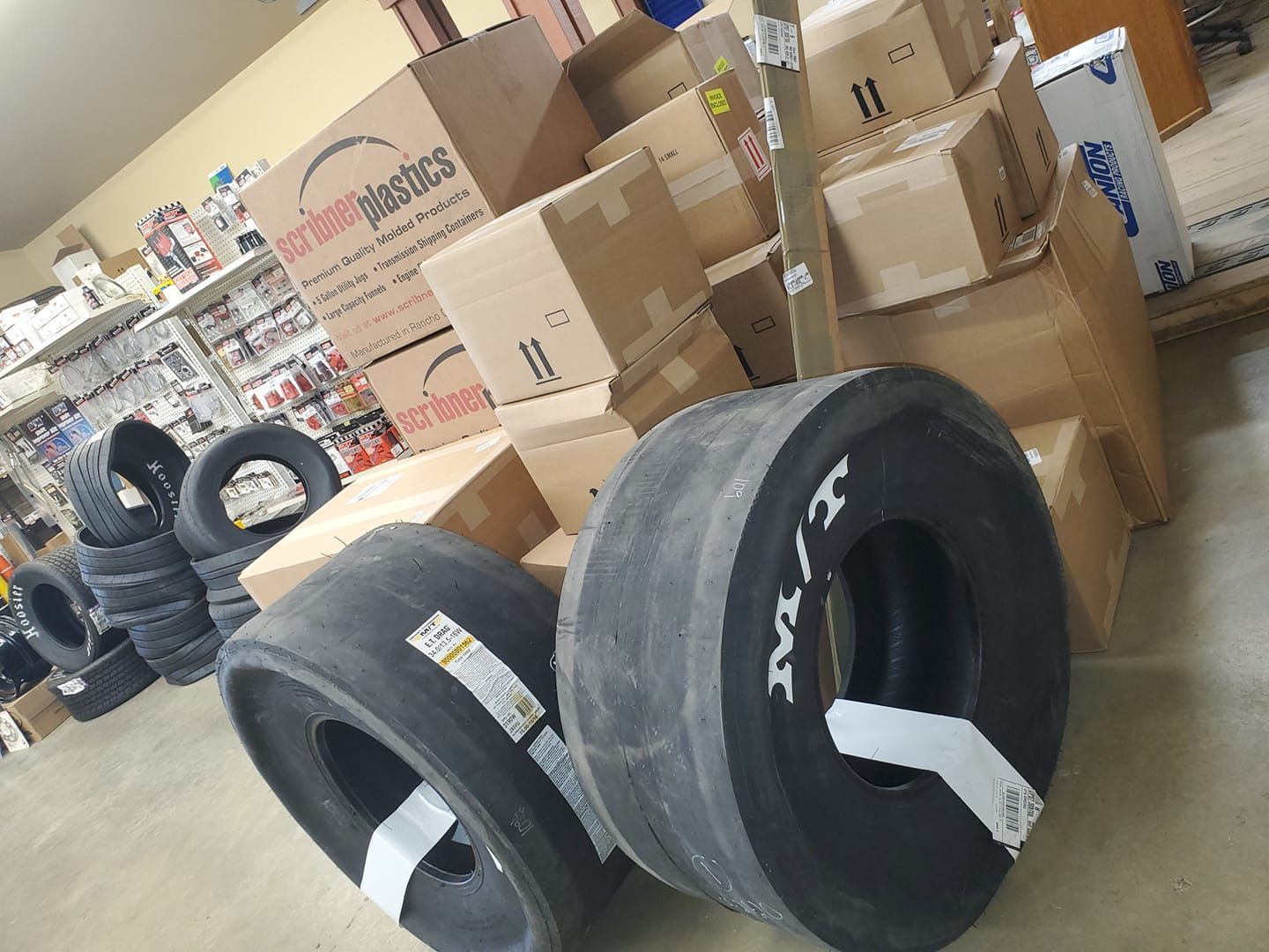 Two racing tires in front of stacked cardboard boxes, tires, and shelves in a storage area.