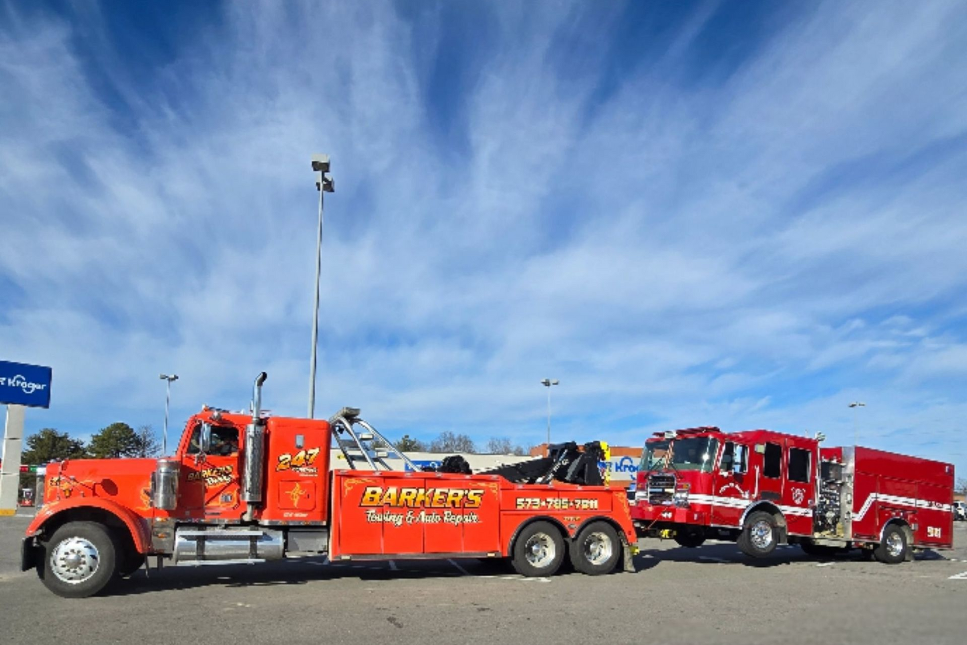 Orange tow truck towing a red fire truck in a parking lot on a sunny day.