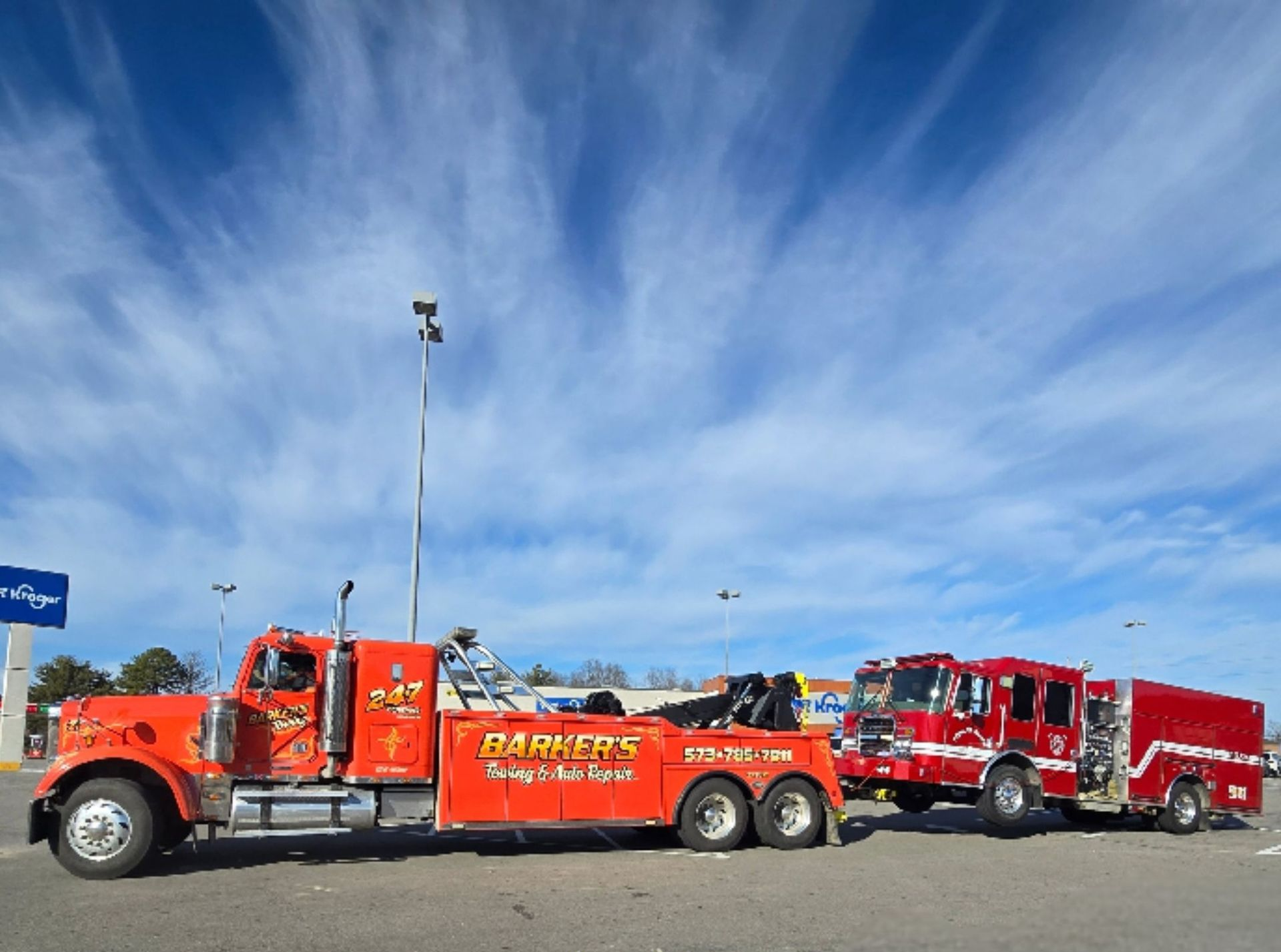Orange tow truck towing a red fire truck under a partly cloudy blue sky.