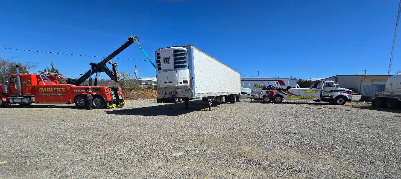 A large orange tow truck lifts a refrigerated trailer on a gravel lot.