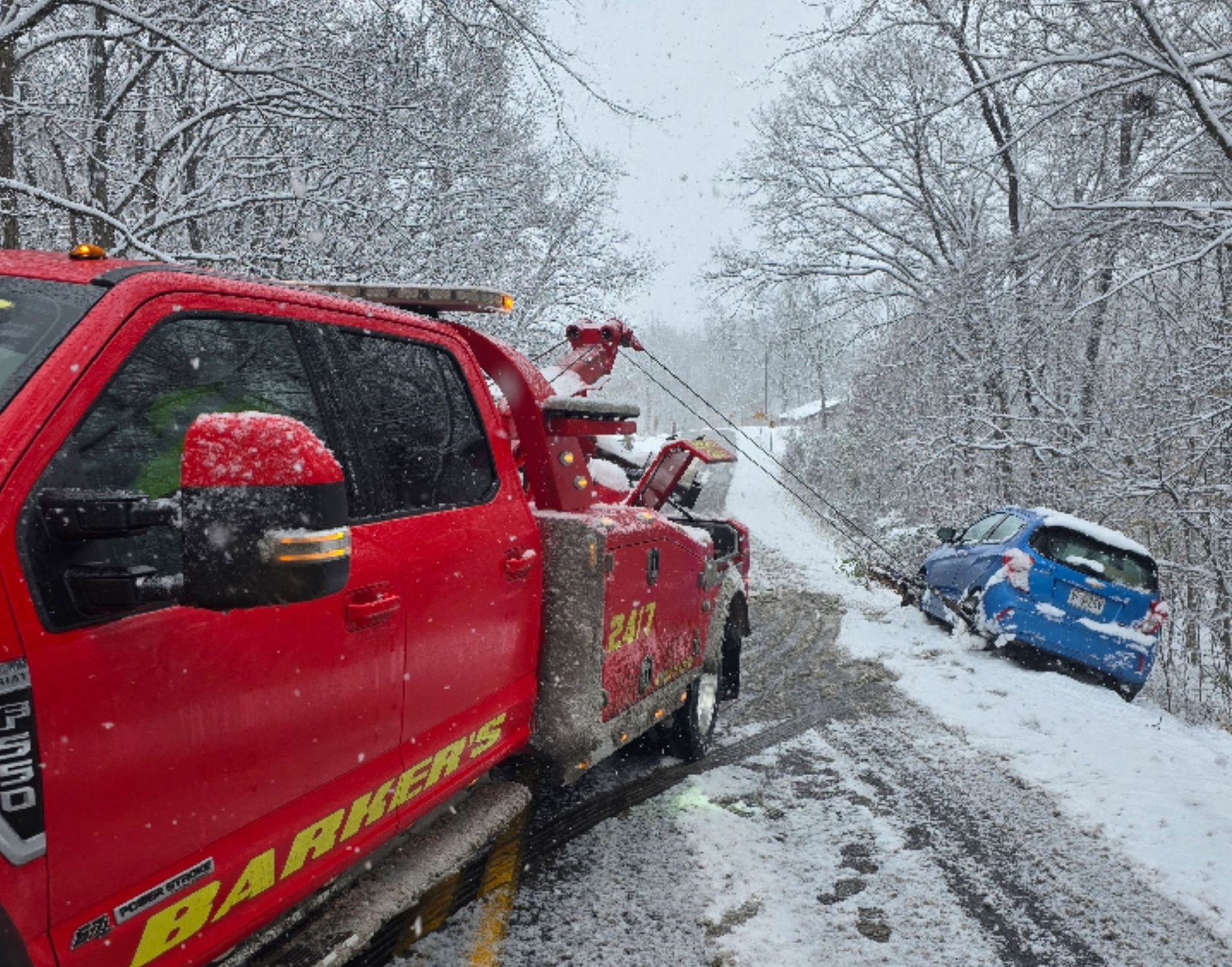 Red tow truck pulling a blue car from a snow-covered ditch on a rural road.