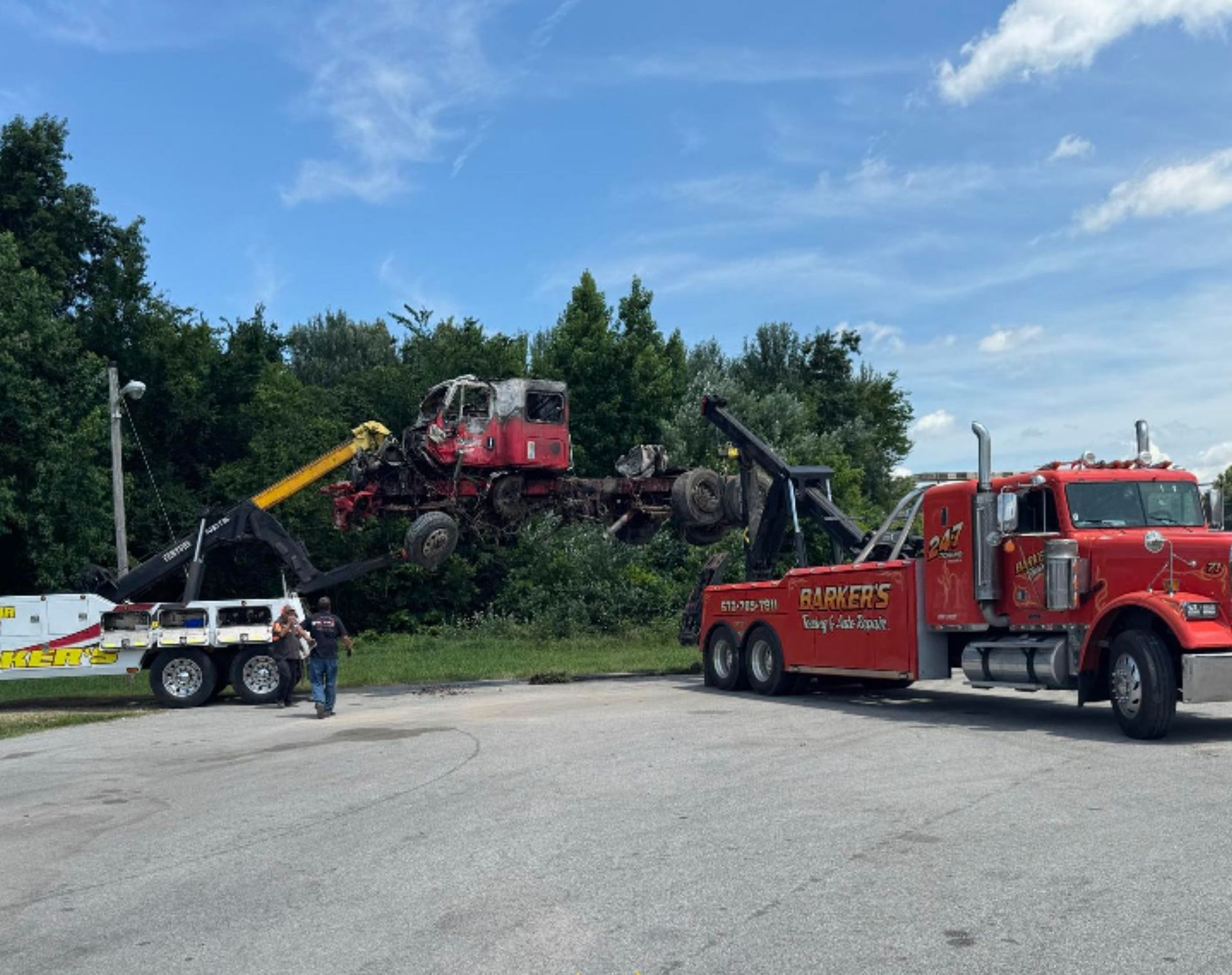 Two tow trucks removing a wrecked red semi-truck from bushes beside a road under a blue sky.