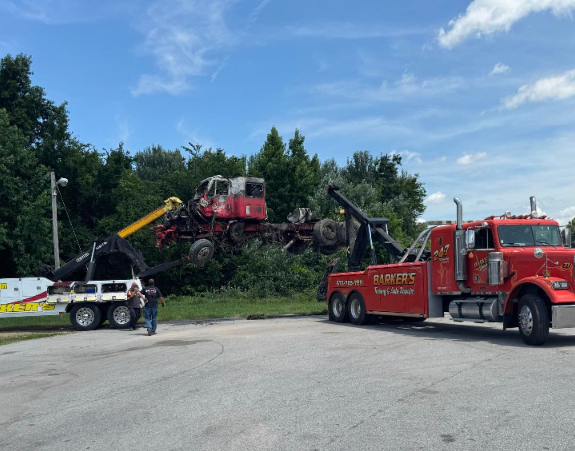 Red tow truck removing a damaged red truck from bushes on a sunny day.