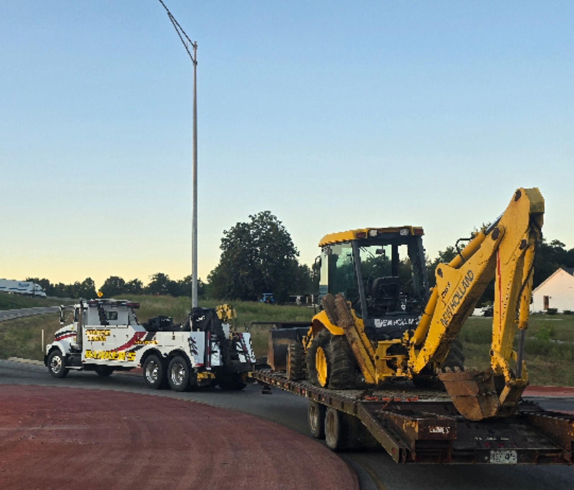 Tow truck pulling a yellow backhoe on a flatbed trailer, likely for transport. Evening setting.