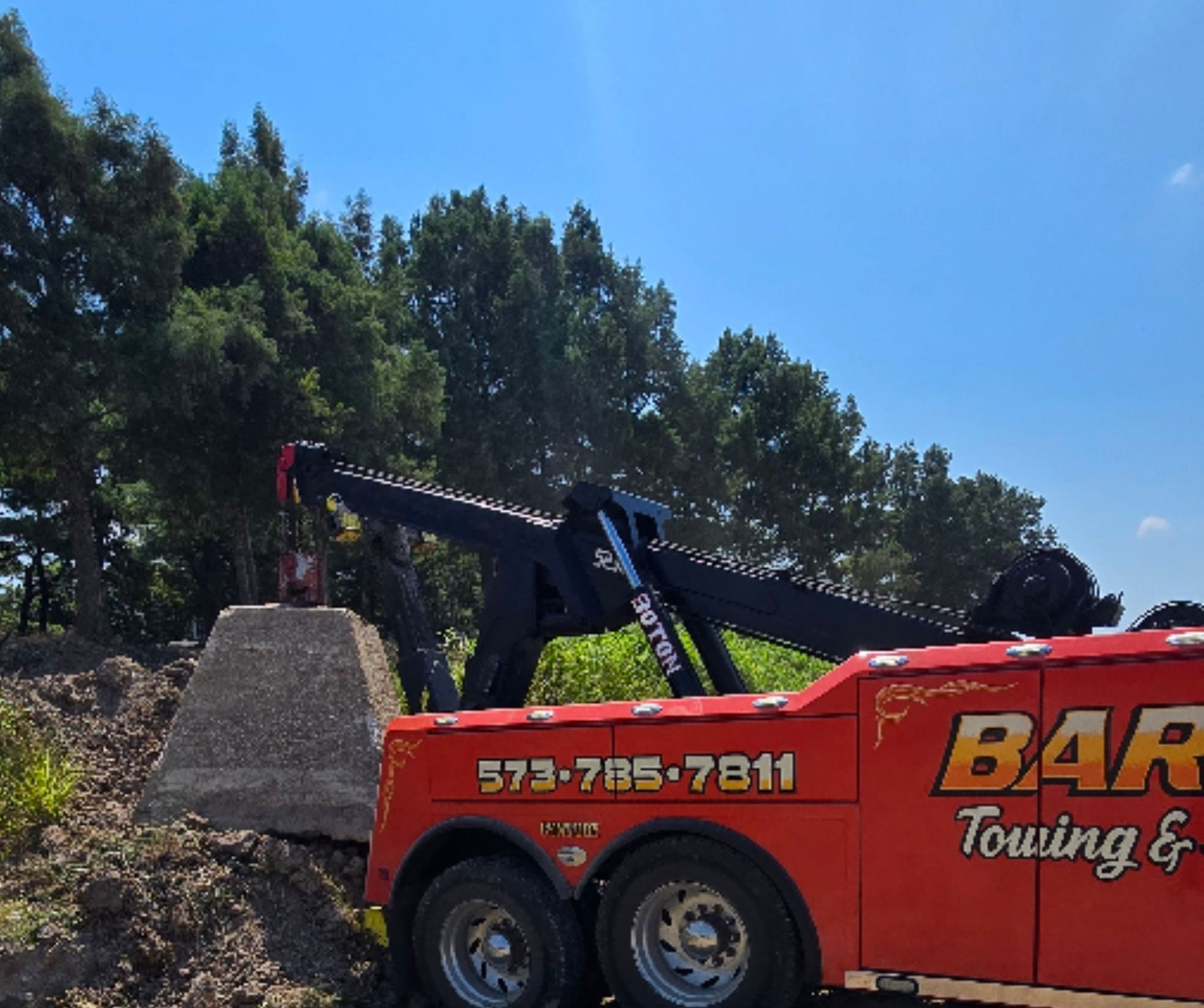Red tow truck lifting a concrete structure outdoors with a phone number on its side.