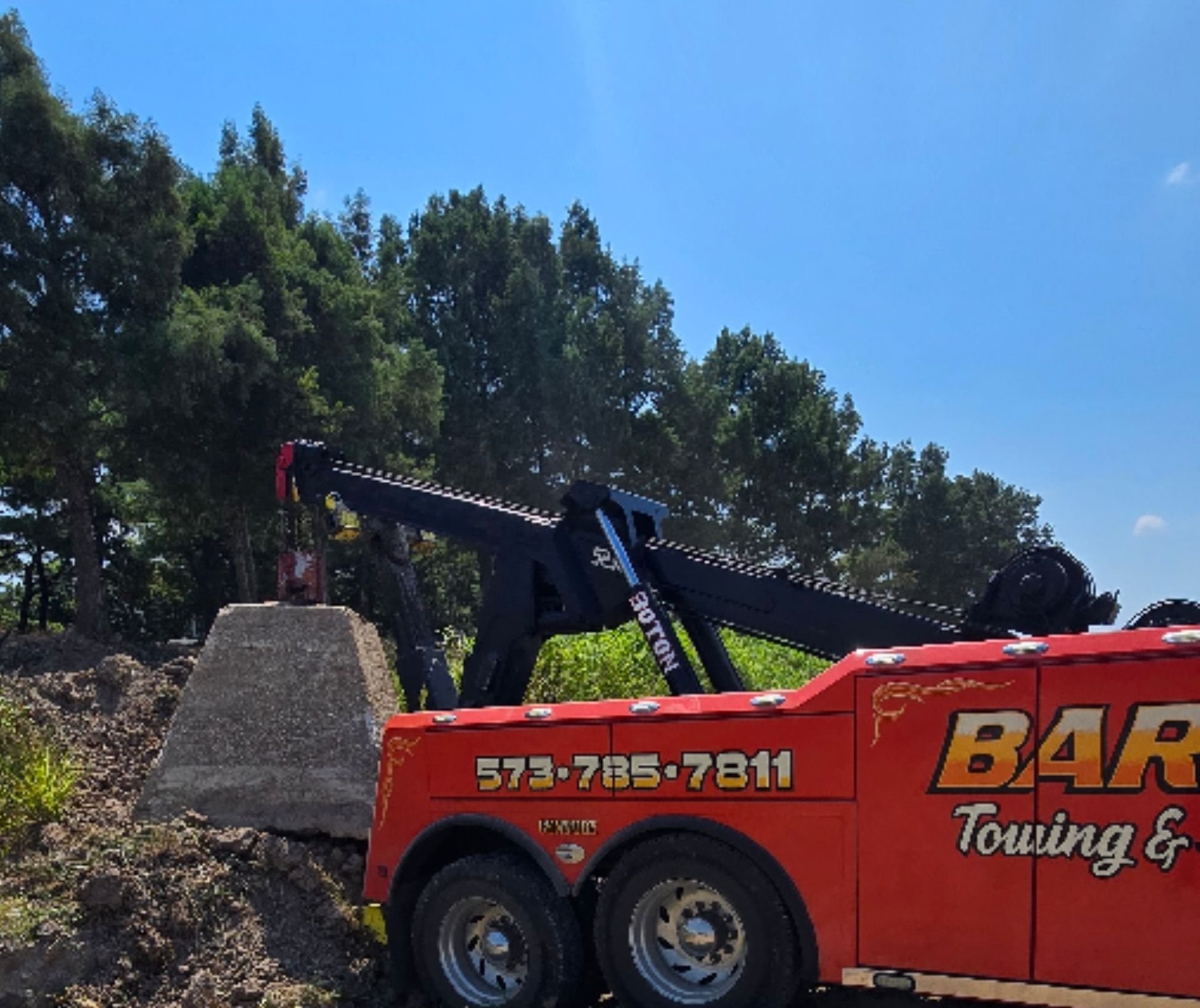 A tow truck lifting a concrete structure, in a rural setting under a blue sky. The truck is orange, and trees in the background.