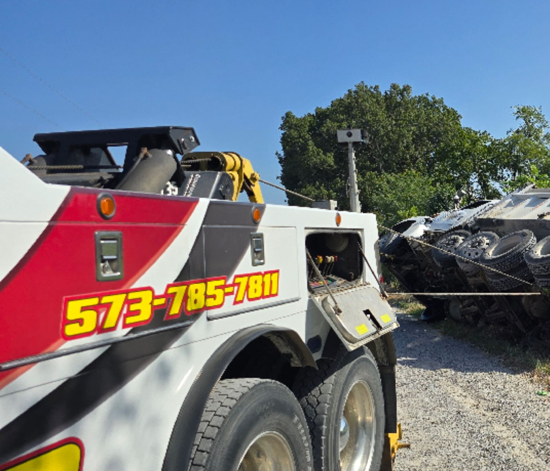 Tow truck with red and white paint, towing a tank on a gravel road, under a blue sky.