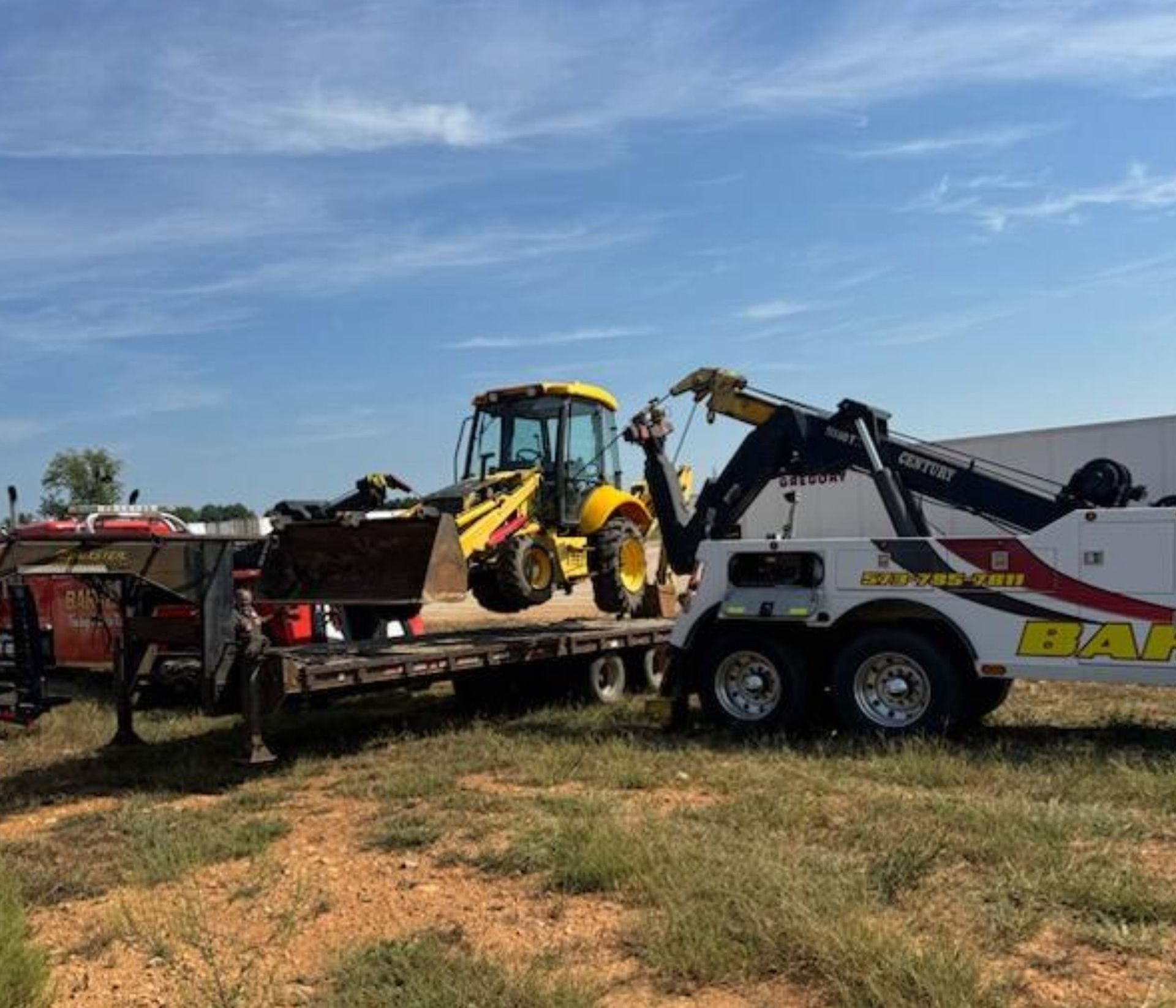 A yellow backhoe being loaded onto a trailer by a tow truck in a field under a blue sky.