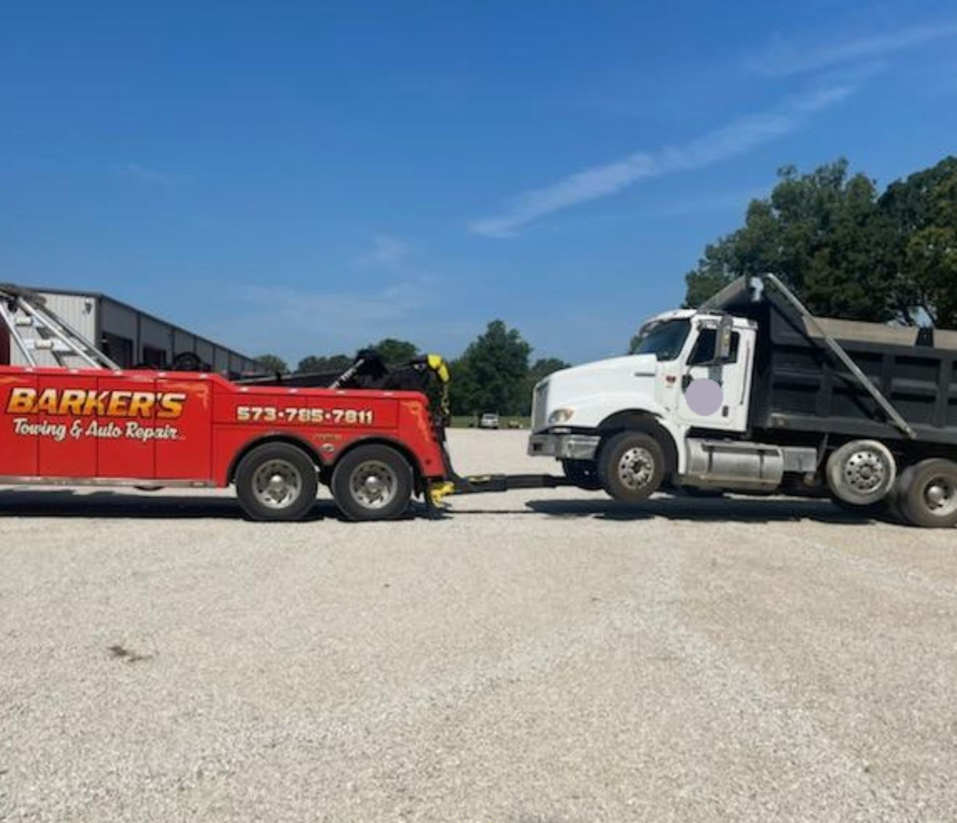 Red tow truck towing a white dump truck on a gravel lot under a blue sky.