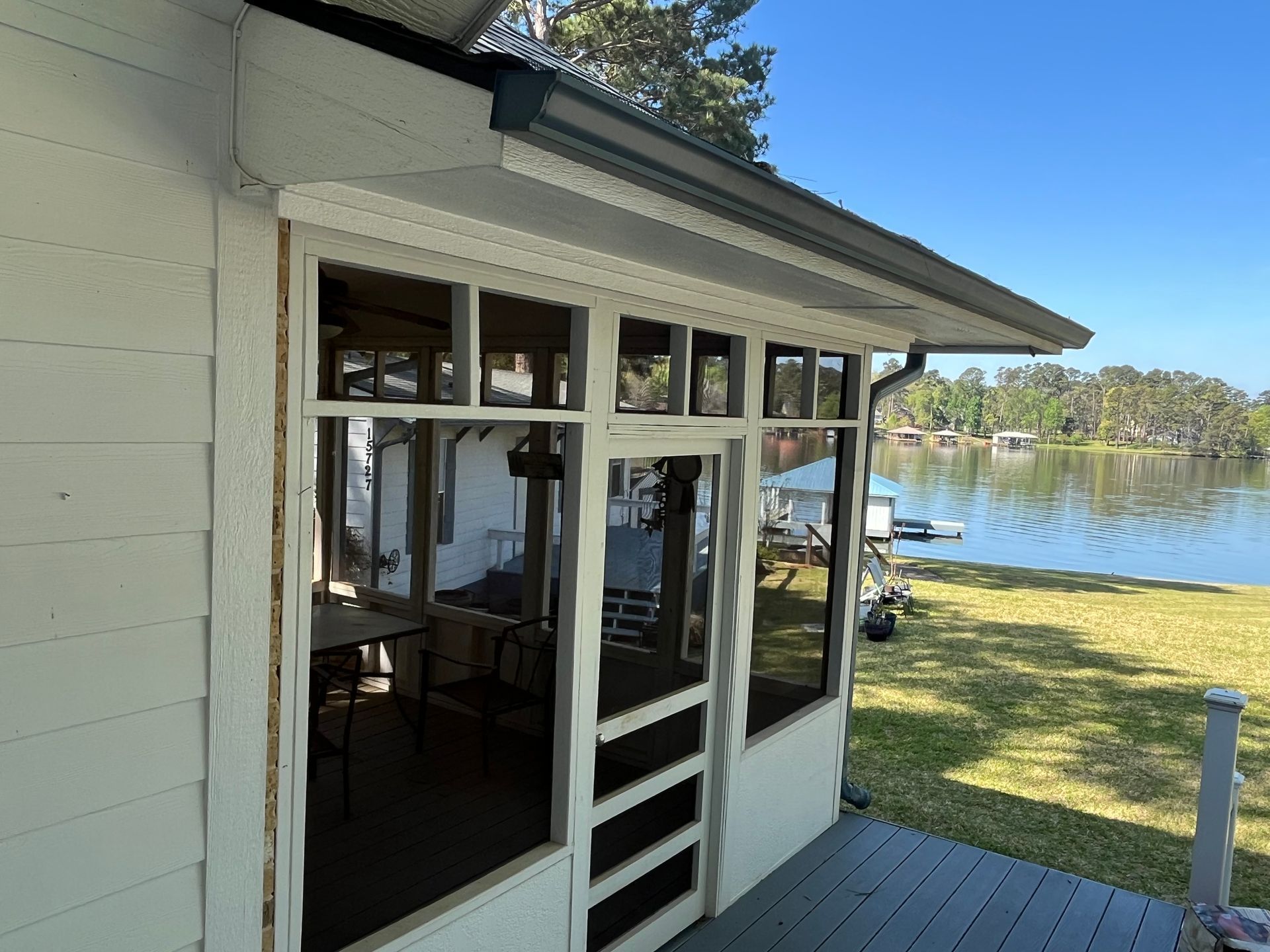 A screened-in porch with white wooden framing overlooks a grassy lawn and a calm lake under a clear blue sky.