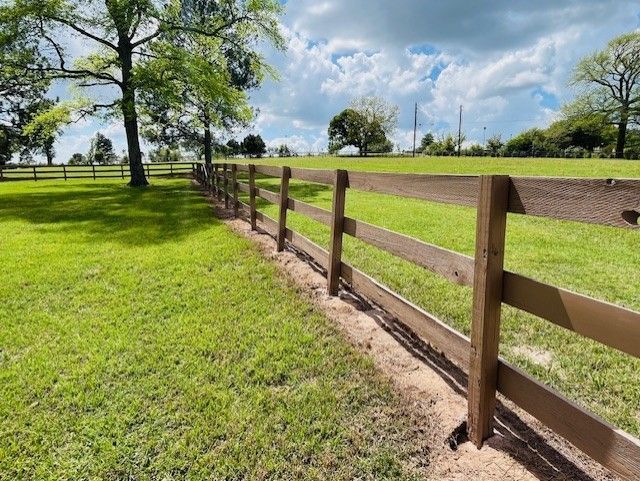 A wooden three-rail fence runs through a grassy field under a bright blue sky with scattered white clouds.