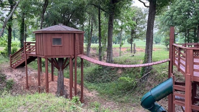 Wooden treehouse on stilts connected by a rope bridge in a wooded park with a slide nearby