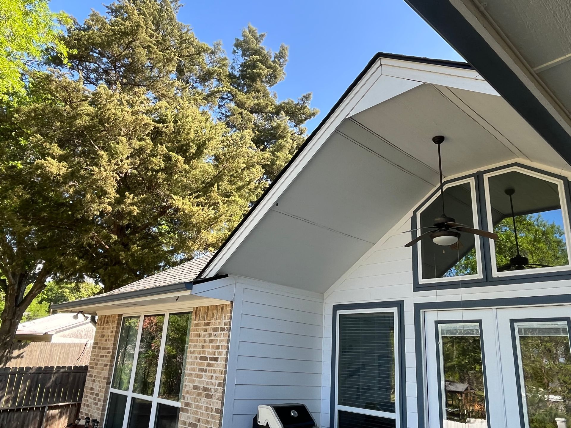 A house exterior featuring a brick wall, white siding, a peaked porch ceiling with a fan, and large windows.
