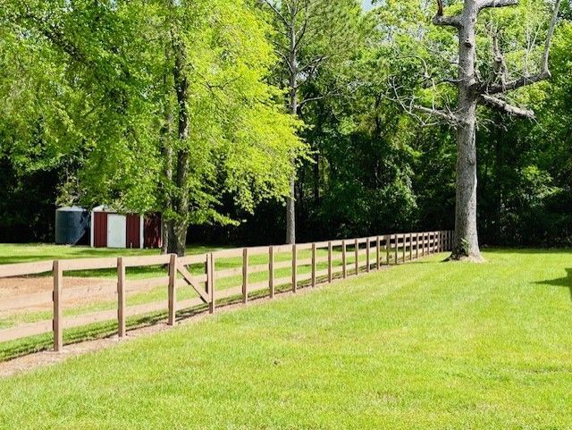 A wooden fence stretches across a green lawn toward a small red shed and trees on a sunny day.