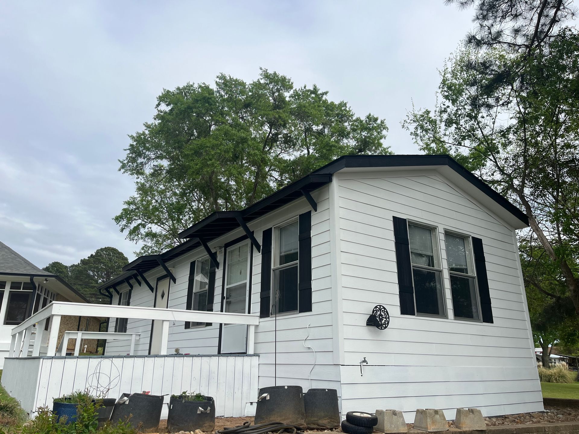 A white mobile home with black window shutters and a small, white wooden deck, situated outdoors near trees.