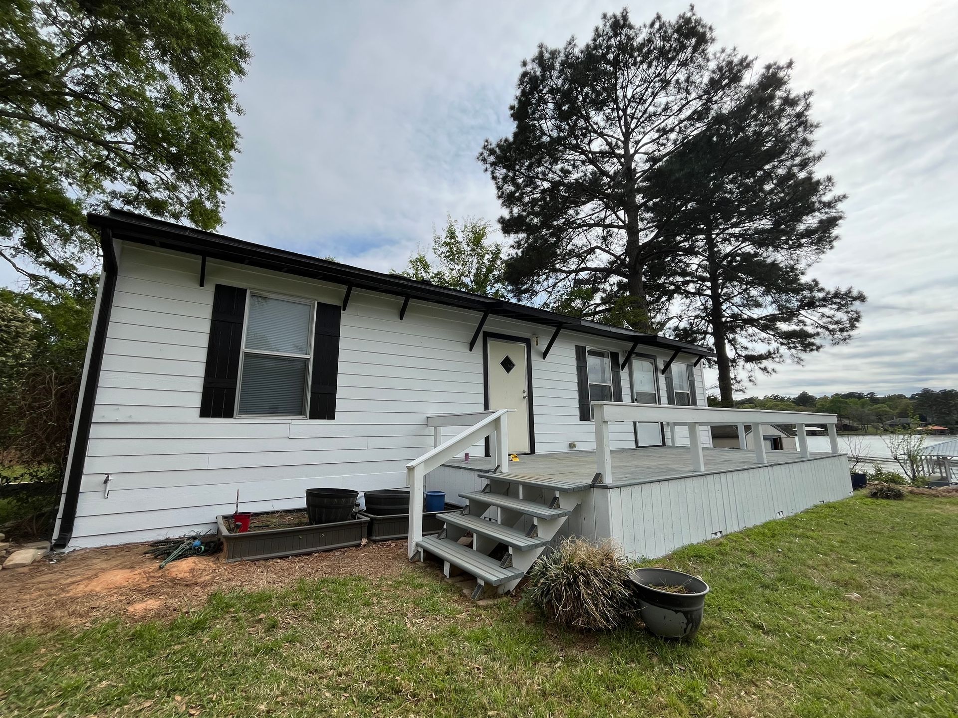 A white, single-story house with black trim and shutters stands beside a deck, overlooking water under a cloudy sky.