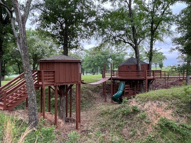 Wooden treehouse play structures linked by a rope bridge in a wooded park, with a slide and stairs