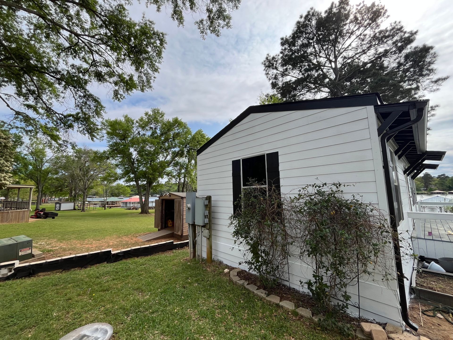 A white house with black trim and shutters stands in a grassy yard, with a small shed and trees in the background.