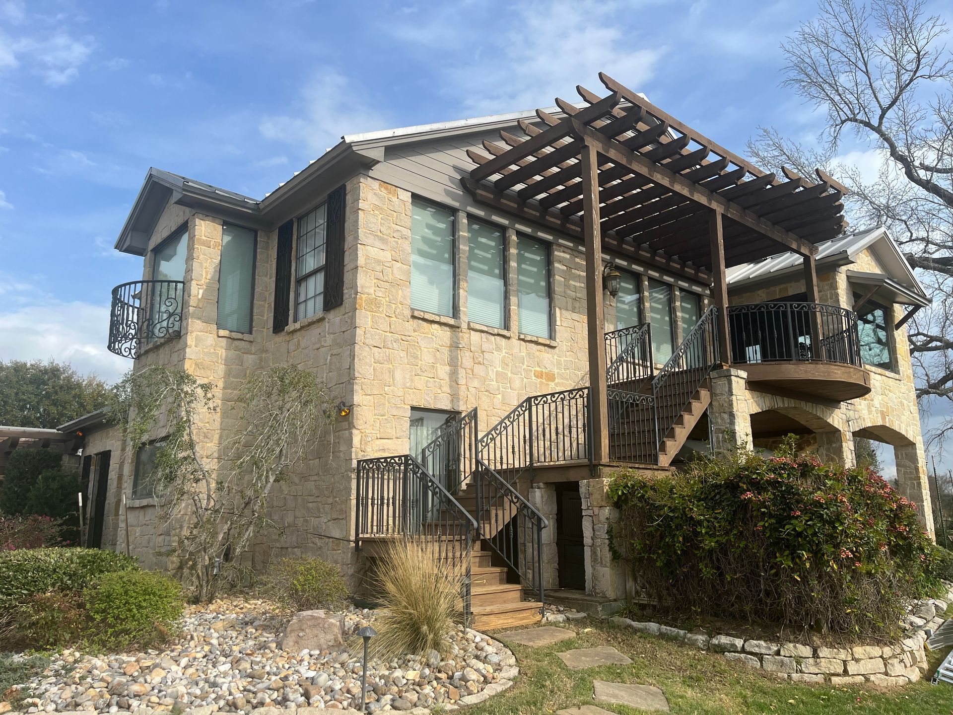 A two-story stone house with a wooden pergola, an outdoor staircase, and a wrought-iron balcony under a blue sky.