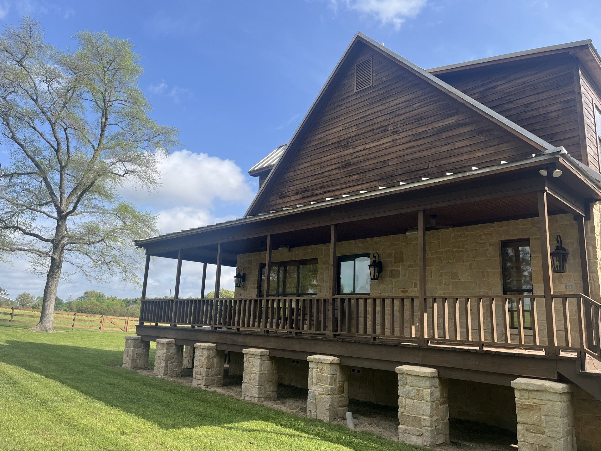 A two-story house with a wooden upper level and stone base, featuring a large wraparound porch next to a tall tree. 