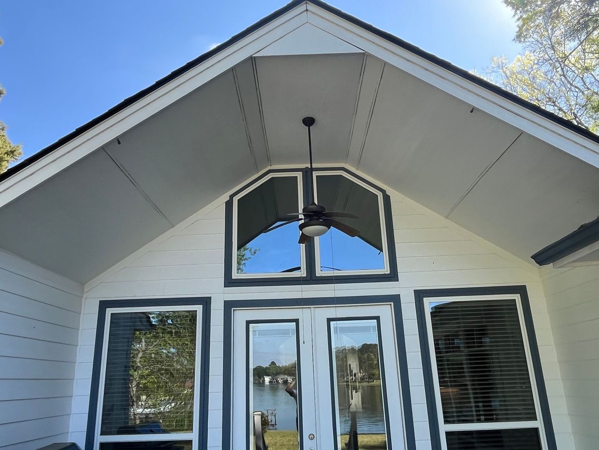 The front exterior of a white-sided house with a vaulted entryway, dark trim, a ceiling fan, and large glass windows.
