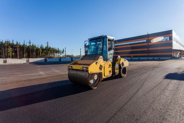 A yellow steamroller compacts fresh asphalt on a sunny day in front of a large industrial building.