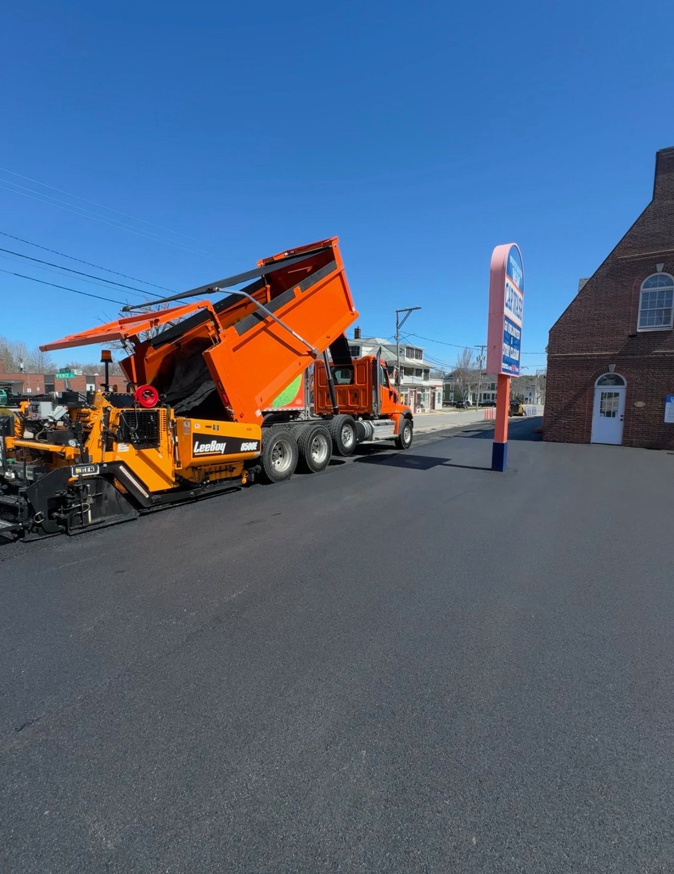 Orange snowplow truck on a sunny street beside a brick building and blue sky
