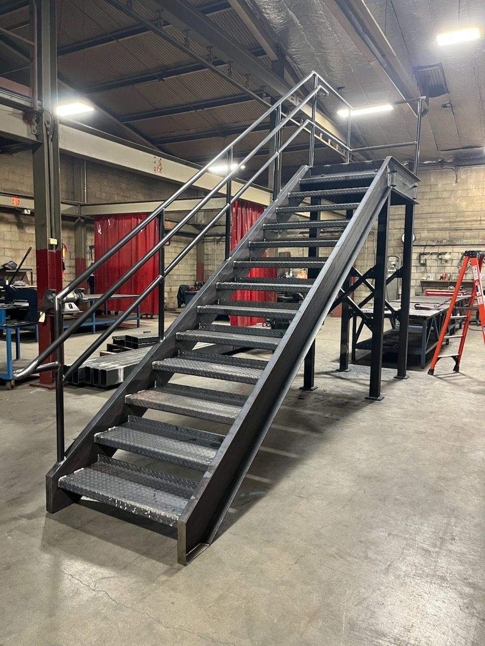 Metal staircase with handrails in an industrial setting. Grey stairs, black supports, and red wall in background.