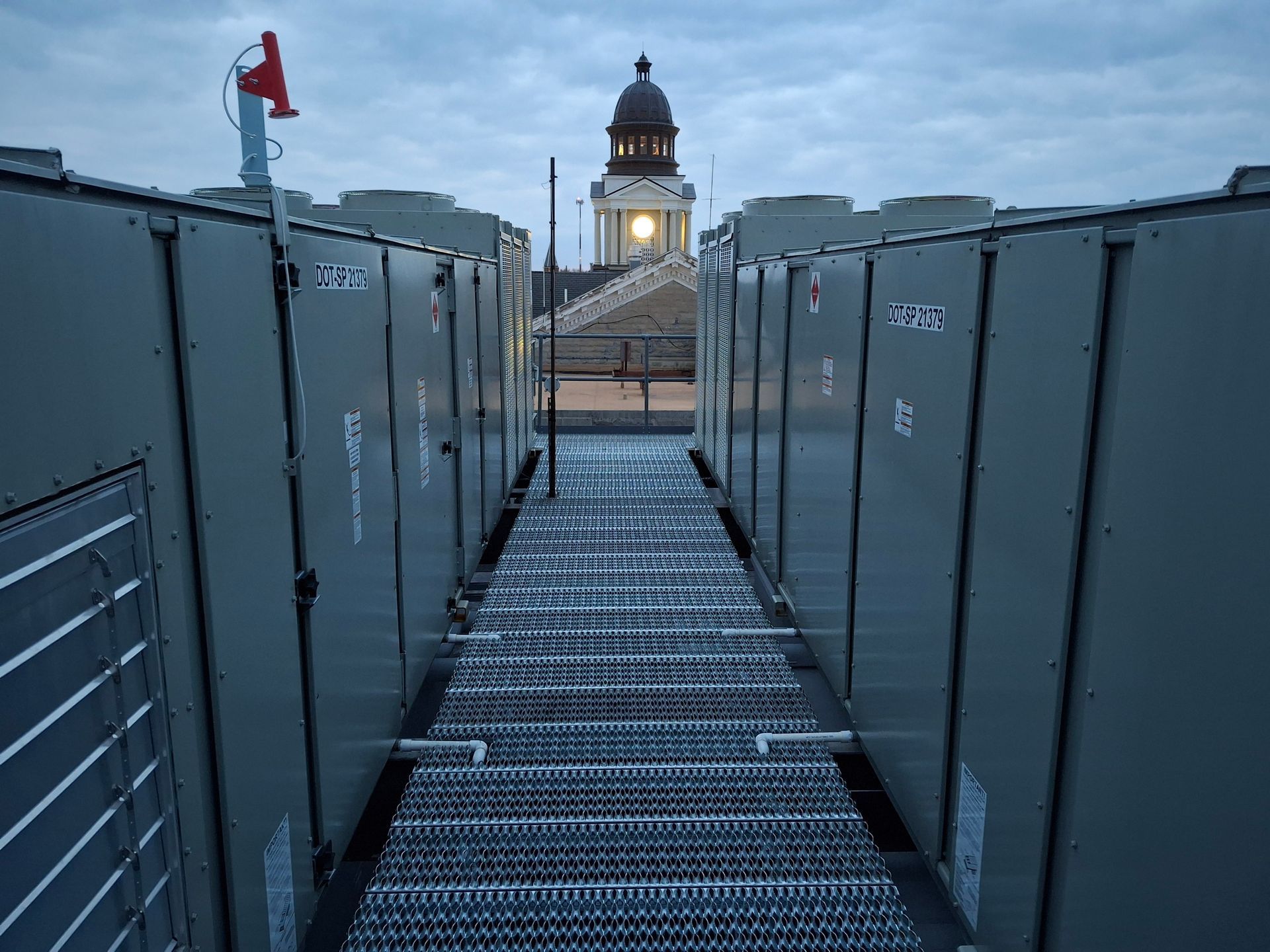 Rooftop with rows of machinery, metal walkway, and a distant building with a dome at dusk.