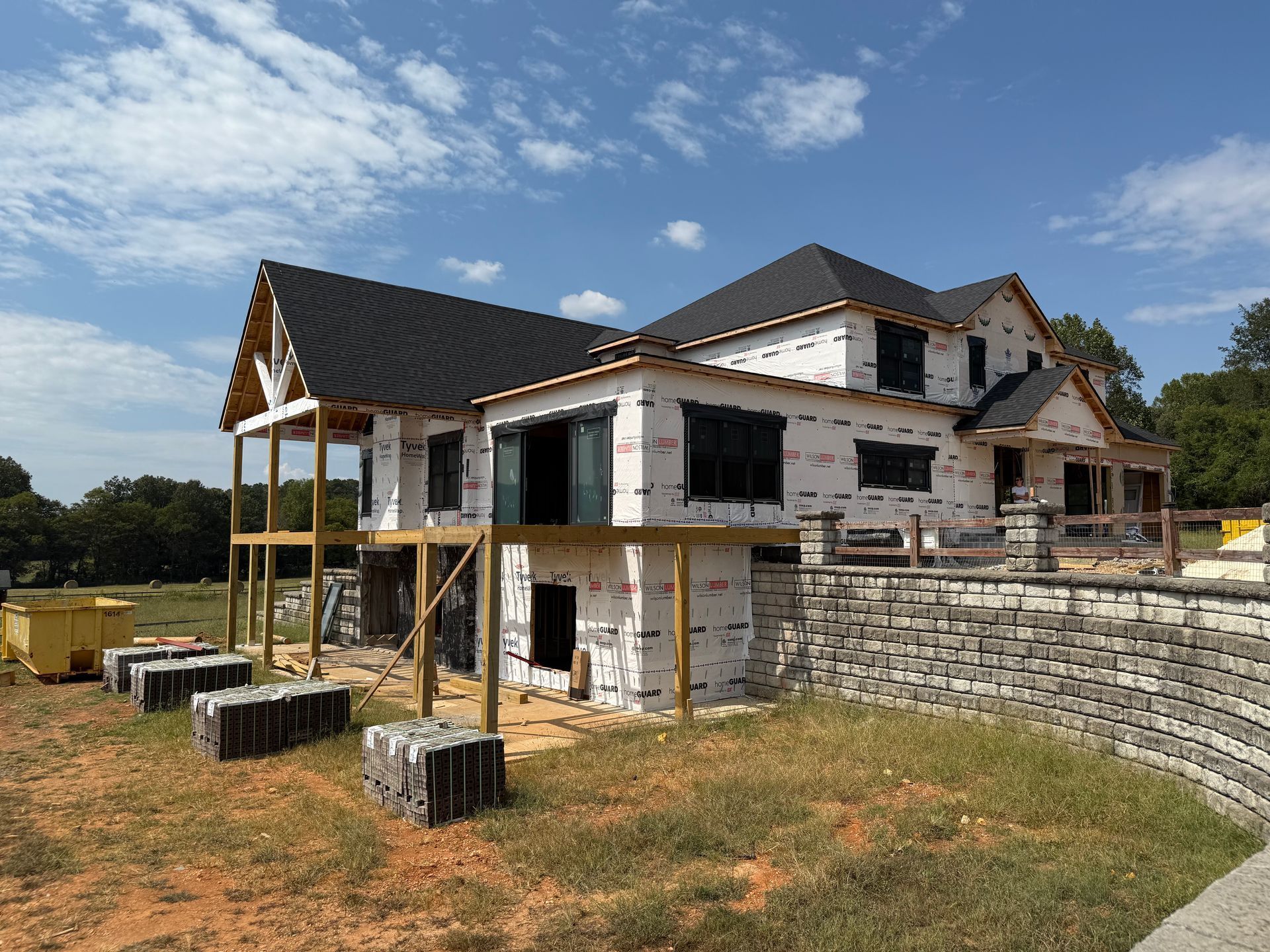 A two-story house under construction with a stone retaining wall and a partially built deck on a sunny day.