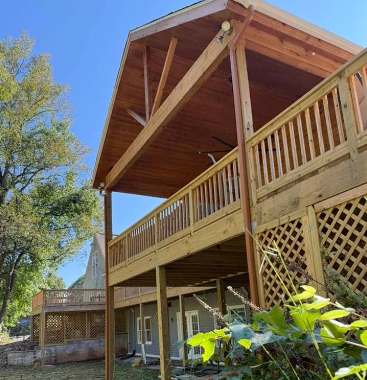 Wooden two-story deck with a covered upper level. Brown ceiling, light wood railings, and lattice panels. Blue sky background.