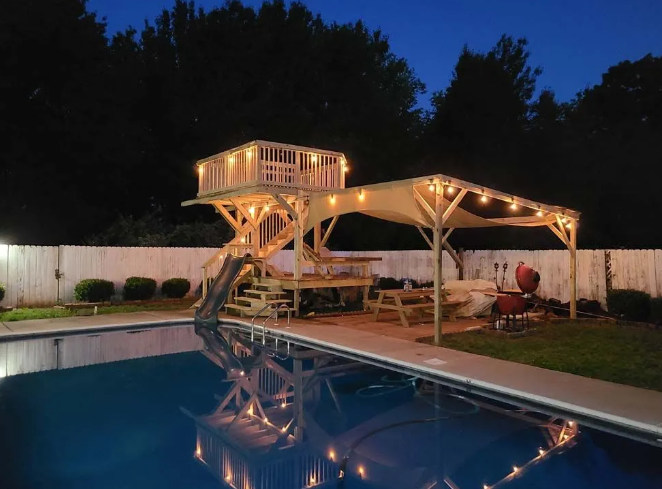 Poolside wooden structure with a slide, deck, and gazebo, lit with string lights at dusk.