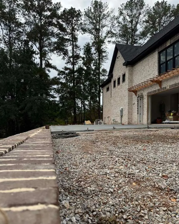 Brick wall and gravel in front of a light brick house with dark roof, trees in the background.