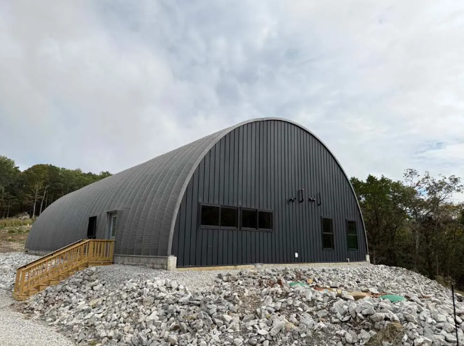 Dark gray Quonset hut building with windows and a wooden staircase. Situated on a rocky hillside under cloudy skies.