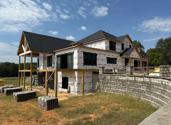 House under construction, covered in white wrap, black roof, and retaining wall.